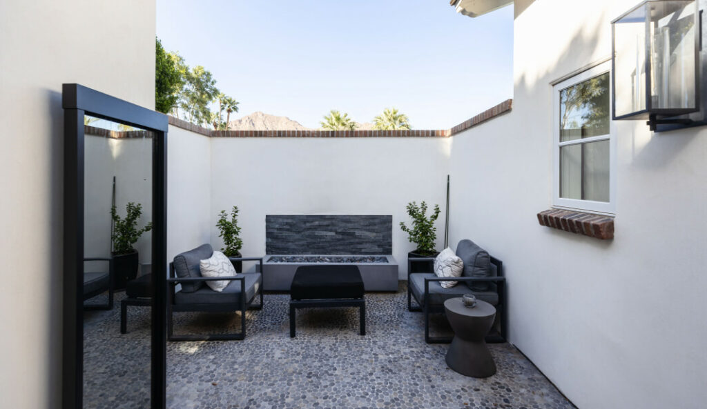 Modern outdoor patio with dark gray armchairs, fire pit, stacked stone wall, pebble flooring, and desert mountain views.