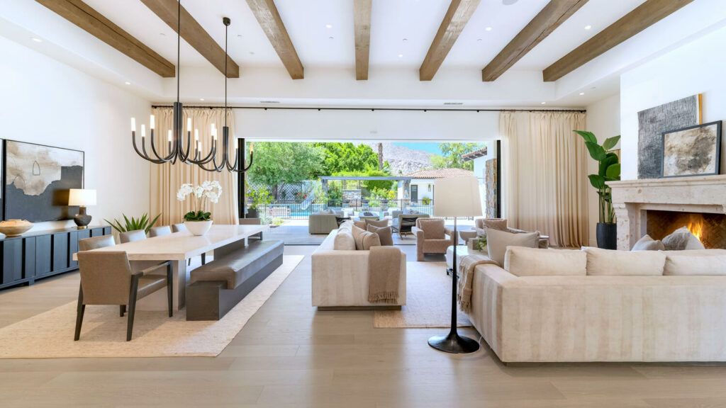 Luxurious open-plan living room with wood beams, contemporary chandeliers, fireplace, and sliding doors to a terrace with a pool, designed by Woodcrest Design.