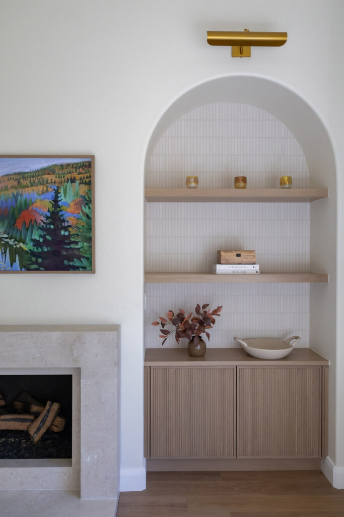 Arched built-in with light wood shelves, fluted cabinetry, patterned tile, gold light fixture, and decorative items. Adjacent modern fireplace and vibrant landscape painting.