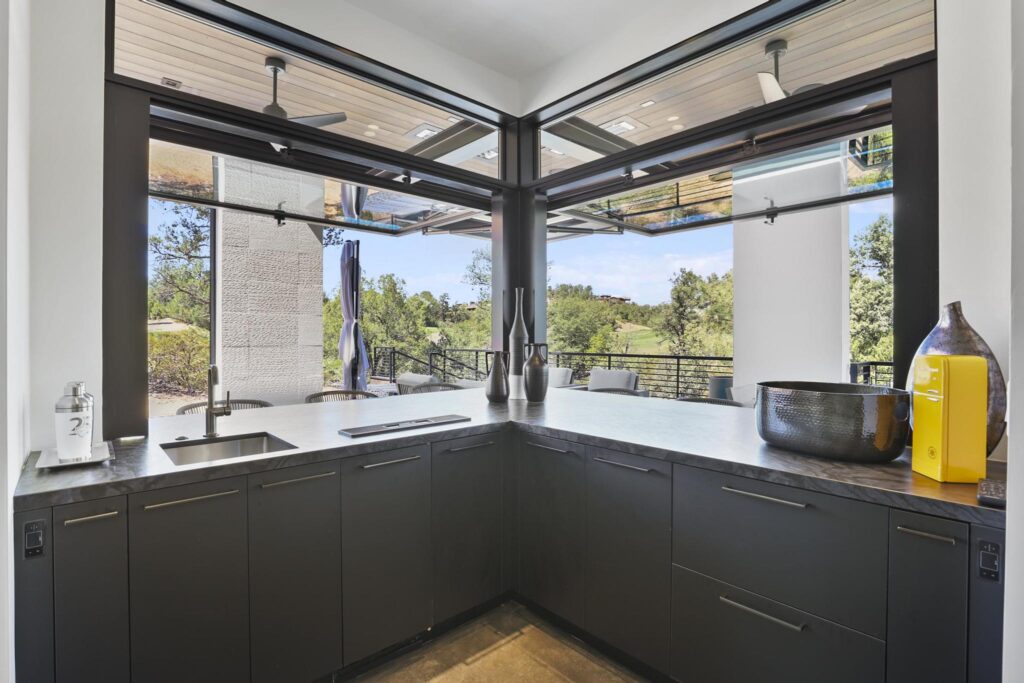 Sleek outdoor kitchen with dark cabinetry, gray countertops, and stainless steel sink. Sliding glass walls open to a green golf course view, featuring decorative vases, a cocktail shaker, and a yellow retro mini-fridge.