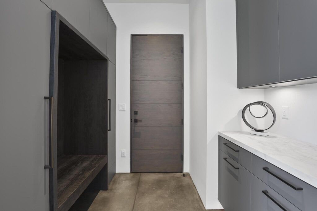 Luxurious modern mudroom with gray custom cabinetry, a built-in dark wood bench, and a sleek dark wood plank door. Contemporary storage with minimalist design.