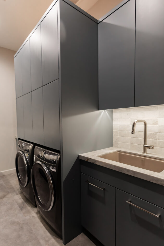 Luxurious laundry room by Woodcrest Design with dark grey custom cabinetry, front-load washer/dryer, sink, and marble countertop.